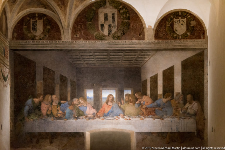 Full view of The Last Supper with Christ and the apostles below three decorated lunettes on the refectory wall.