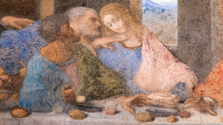 Three apostles lean together in blue, green, and rose garments at the table in Leonardo’s The Last Supper.
