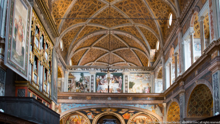 Long view of the former nuns’ hall with fresco-covered walls, upper galleries, arched bays, and an ornate patterned ceiling.