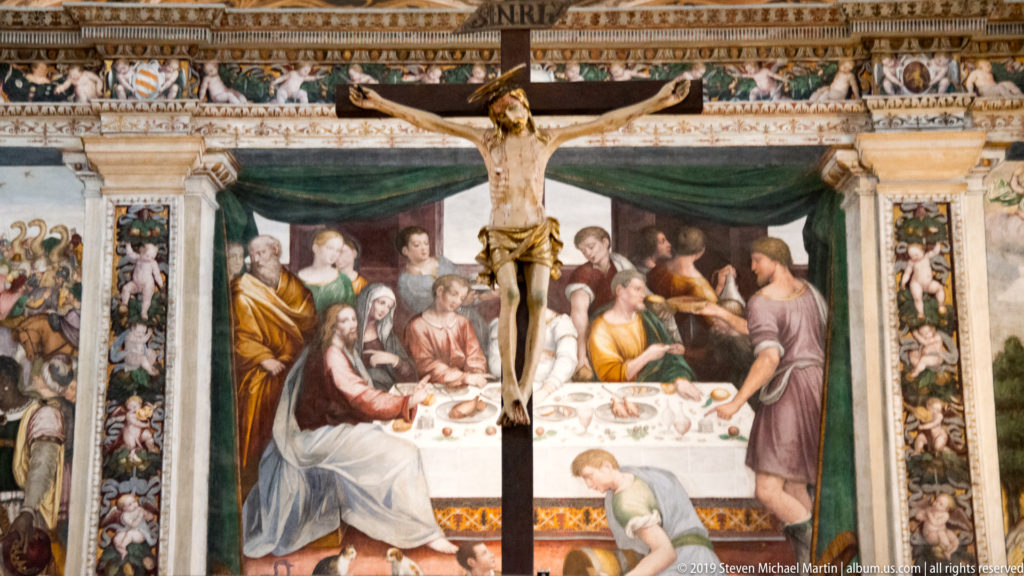 Large crucifix in front of a Last Supper fresco with Christ and apostles seated around a long table.