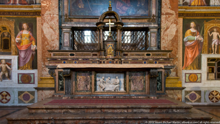 Frontal altar with central tabernacle, metal grille, colored marble, painted saints, and a white Last Supper relief.