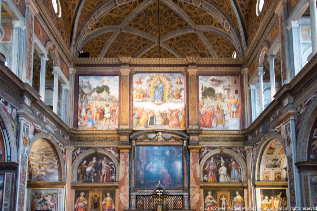 Frontal view of a fresco-covered partition wall with large narrative scenes, framed saints, and a central altar area.