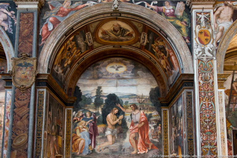 Fresco of a baptism in a landscape, with a nearly nude figure in water, a robed officiant, and attendants beneath an ornate arch.