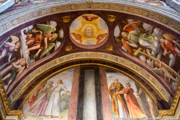 Decorated arch with reclining painted figures and a central roundel above two standing sacred figures in the lunette.