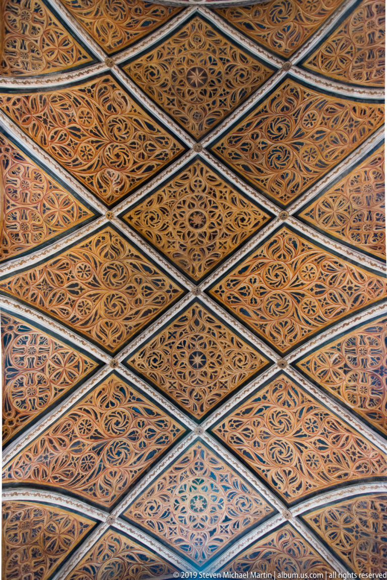 Close view of a painted vaulted ceiling with interlaced diamond compartments filled with intricate foliate ornament.
