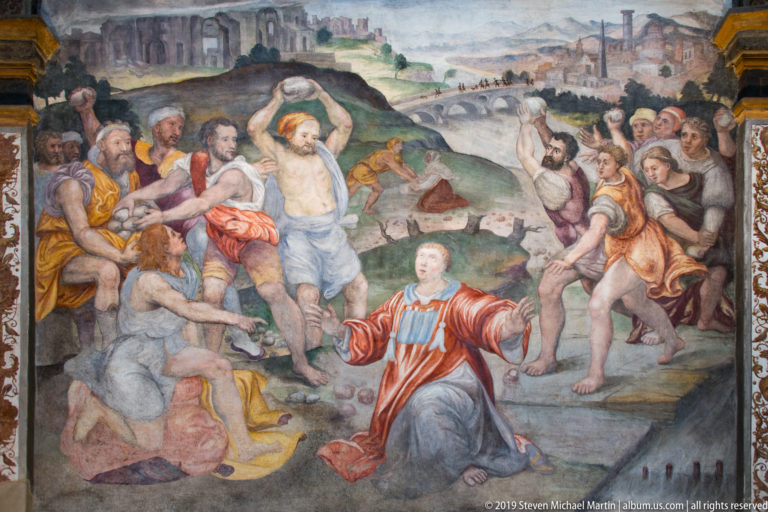 Close view of Saint Maurice kneeling among armed soldiers and executions, with a rocky landscape and distant city behind.