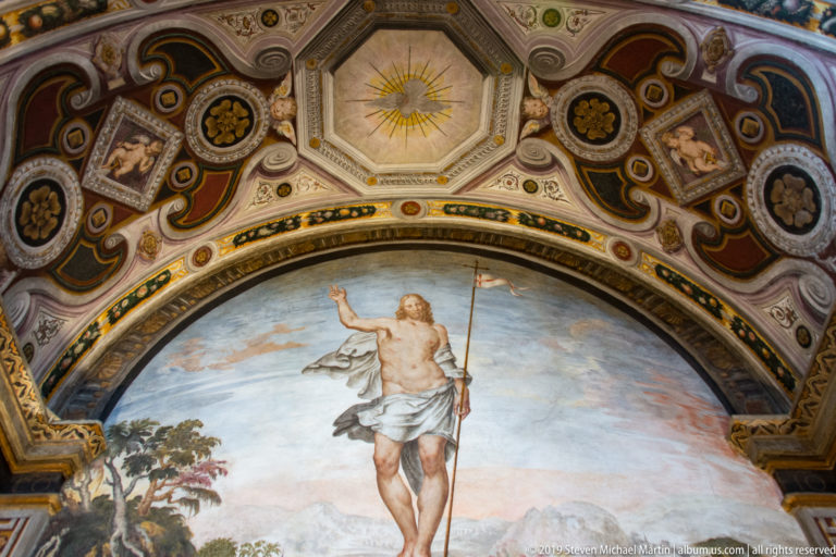 Risen Christ holding a cross-staff in a landscape, framed by an ornate arch with rosettes and painted decorative panels.