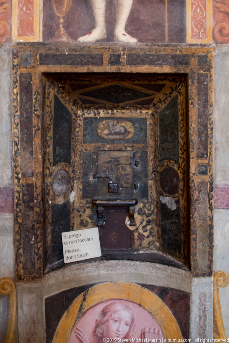 Small rectangular opening with a dark metal grille set into a painted and marbled church wall.