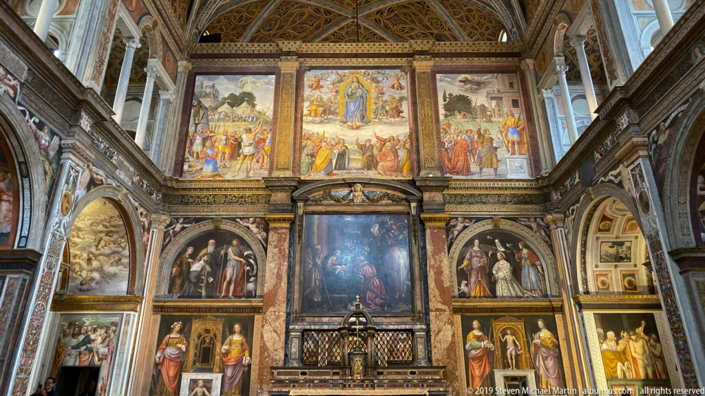 Frontal view of a richly frescoed church partition with altar, grille, saints, and narrative panels arranged in architectural tiers.