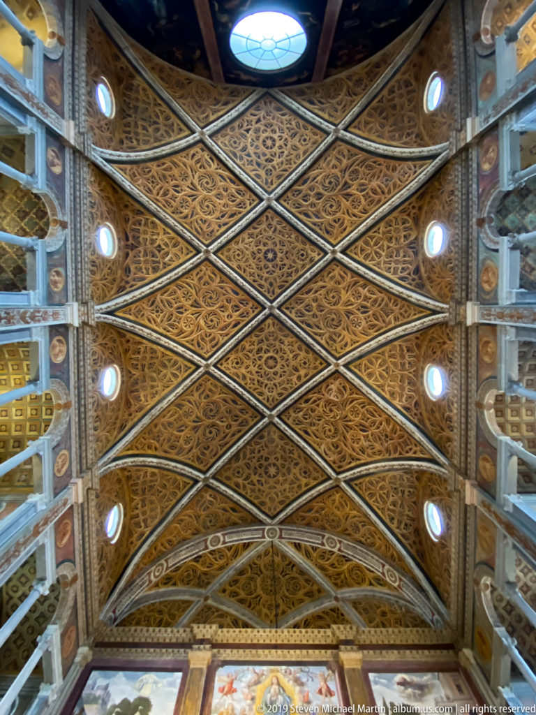 Upward view of a long vaulted ceiling covered in ornate diamond-patterned compartments with circular side windows.