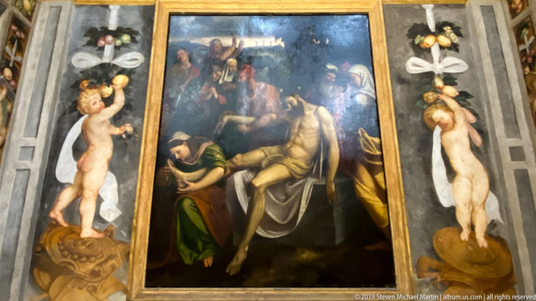 Dark Passion scene with mourners supporting Christ, flanked by painted nude putti holding fruit and floral festoons.