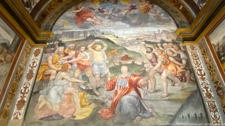 Renaissance fresco of Saint Maurice kneeling amid soldiers and executions in a rocky landscape, framed by ornate painted borders.