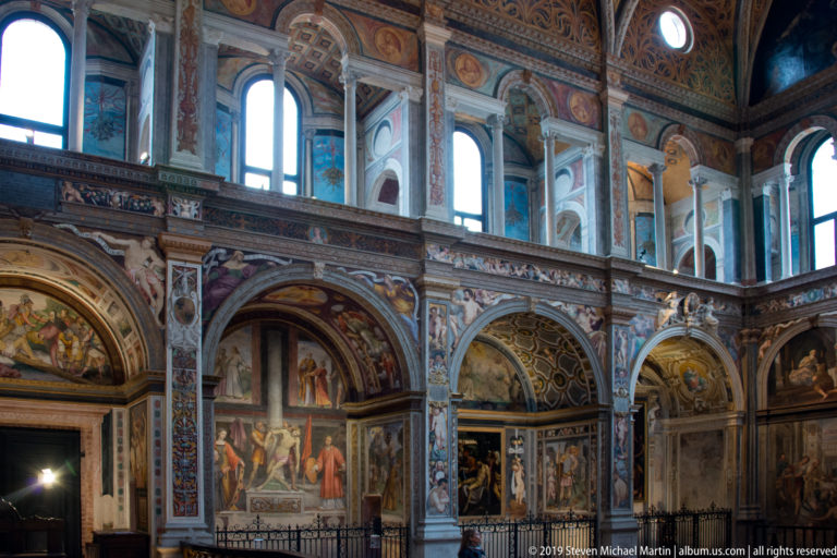 Oblique view of a frescoed side wall with painted arches, narrative scenes, pilasters, and bright clerestory windows.