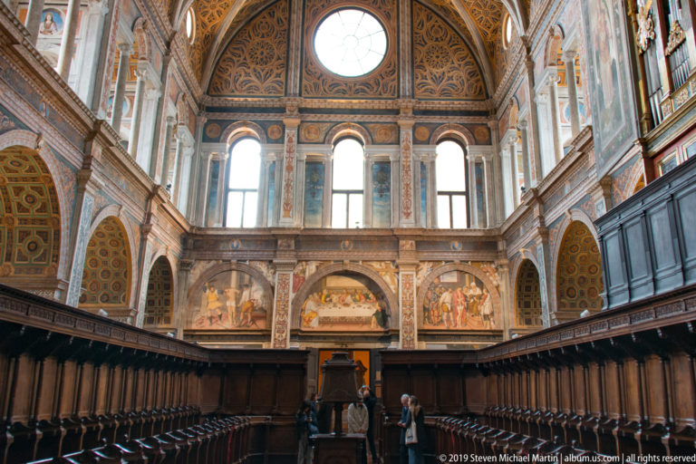 Rows of dark wooden choir stalls below a frescoed gallery with tall windows and a patterned coffered ceiling.