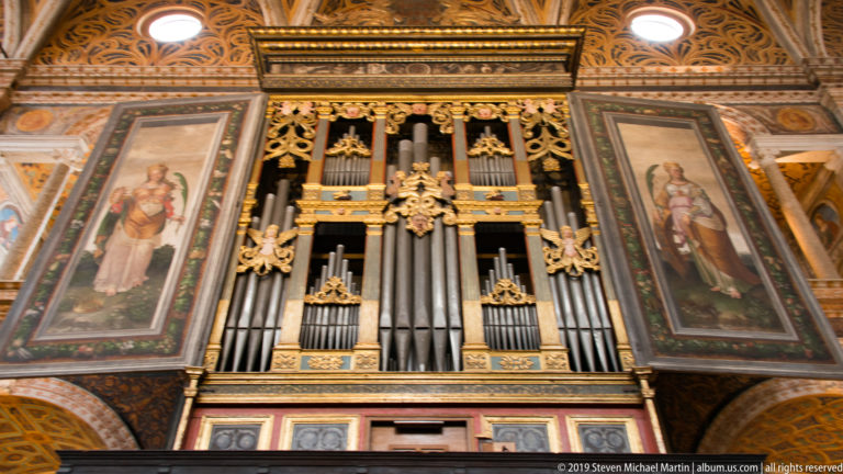 Large pipe organ with gilded case and metal pipes, flanked by open painted shutters showing full-length saints.