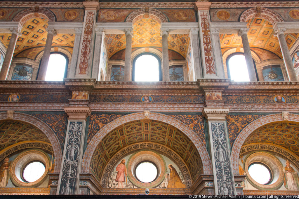 Upper interior wall with tall gallery openings, circular windows, frescoed arches, pilasters, and a richly decorated ceiling.