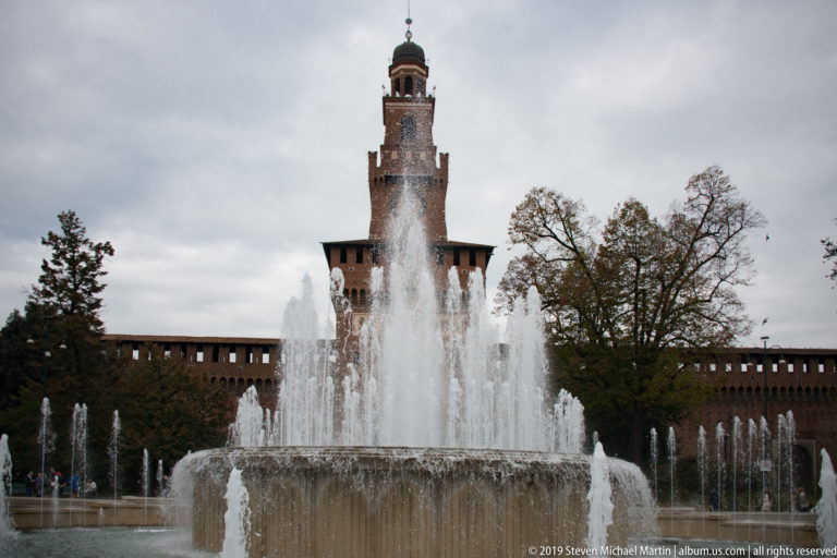Tall fountain jet rising in front of the Torre del Filarete, with smaller jets, trees, and castle walls around it.