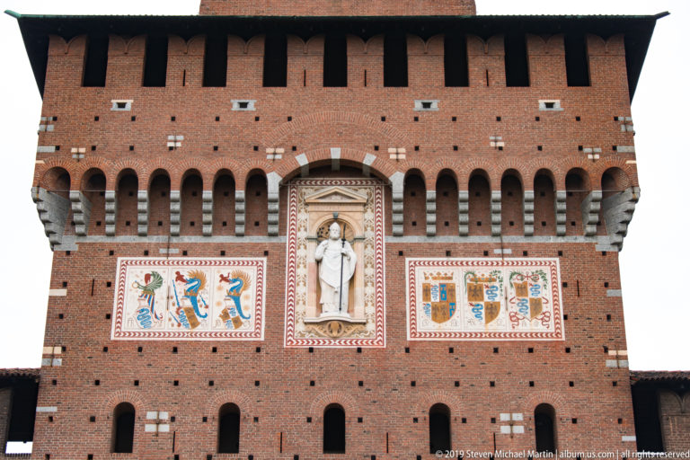 White statue in a tall niche between painted heraldic panels on the red-brick Torre del Filarete façade.