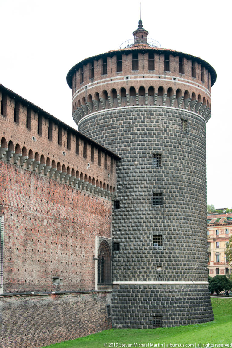 Large round stone-faced tower with brick corbelled crown and small arched doorway at Castello Sforzesco.