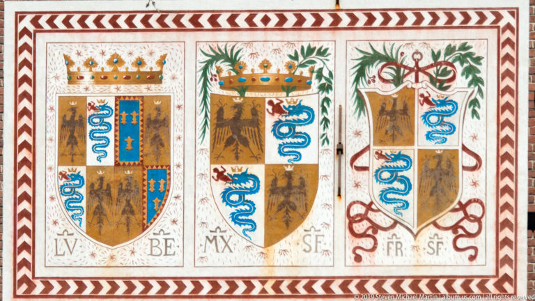 Painted quartered shields with black eagles, blue serpent emblems, crowns, initials, and foliage on a castle façade.