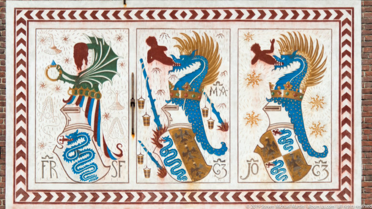 Three painted heraldic devices with eagles, blue serpent emblems, helmets, and initials inside a geometric border.