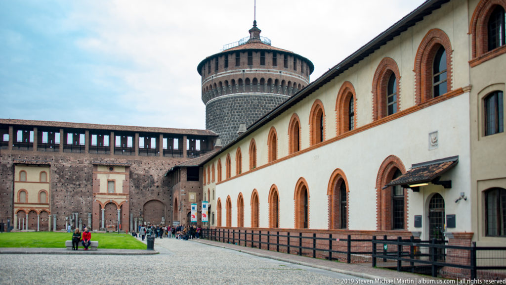 Round stone-faced castle tower behind a long pale building with repeated terracotta-trimmed arched windows at Castello Sforzesco.
