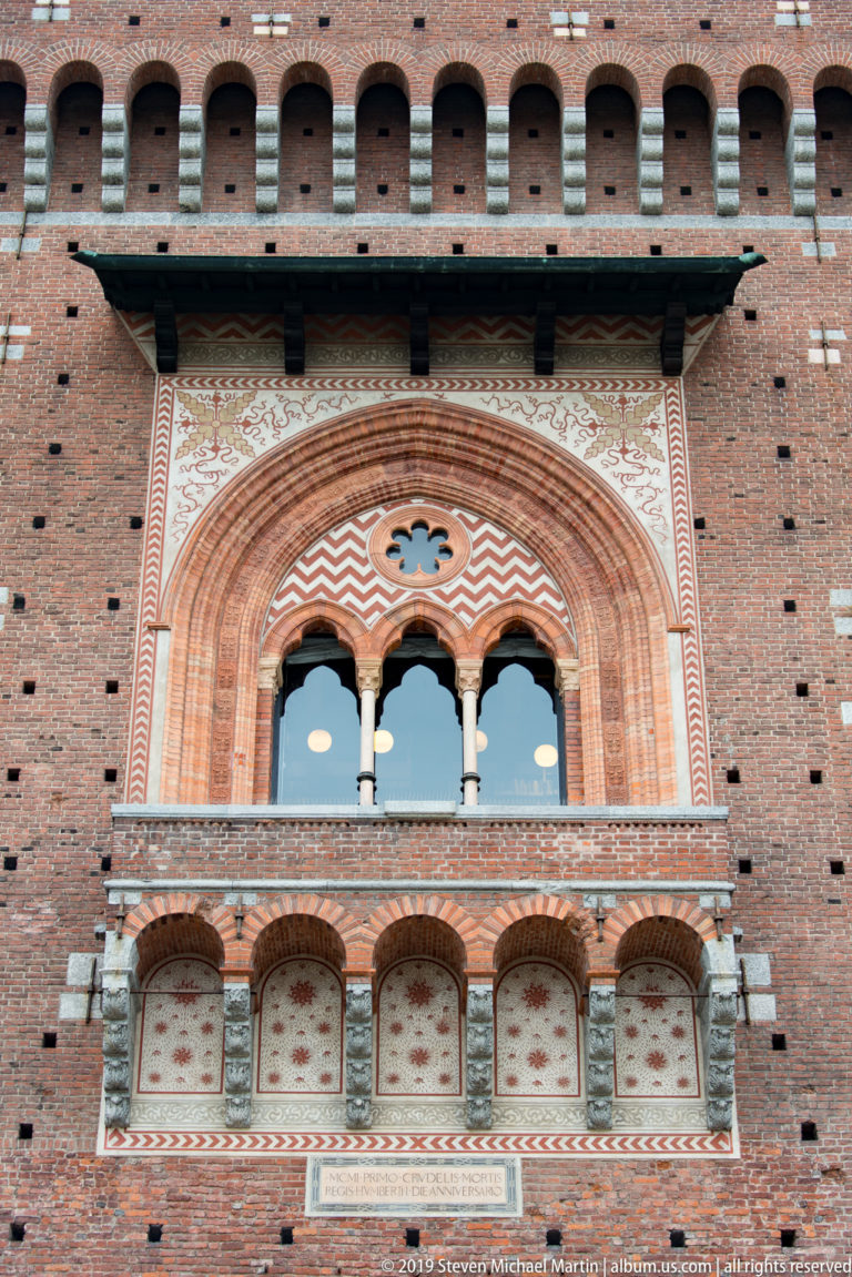 Decorated two-light arched window with central column, patterned brickwork, and painted geometric bands on the Torre del Filarete.