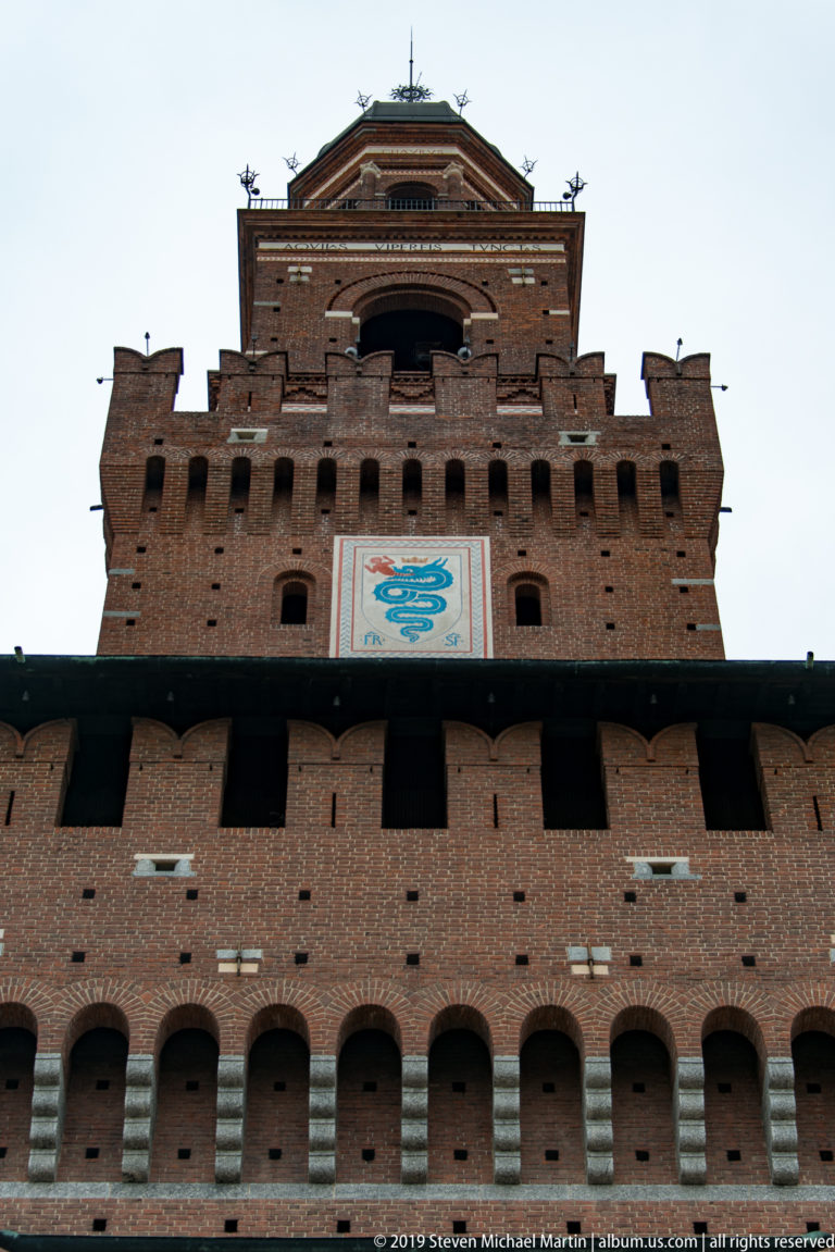 Low-angle view up the brick Torre del Filarete, showing arched openings, projecting corbels, and a large heraldic panel.