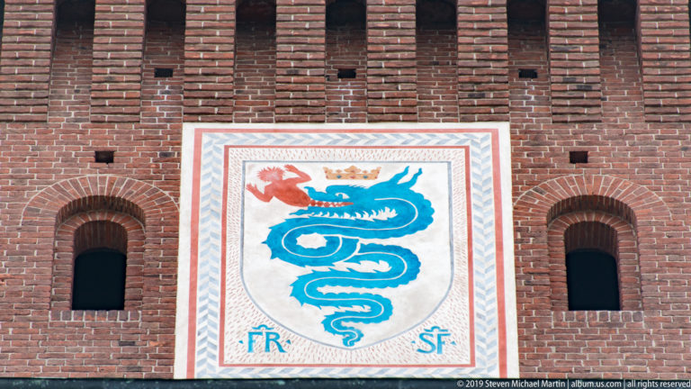 Crowned shield with a blue biscione serpent and the initials FR and SF between two arched openings on a brick tower.
