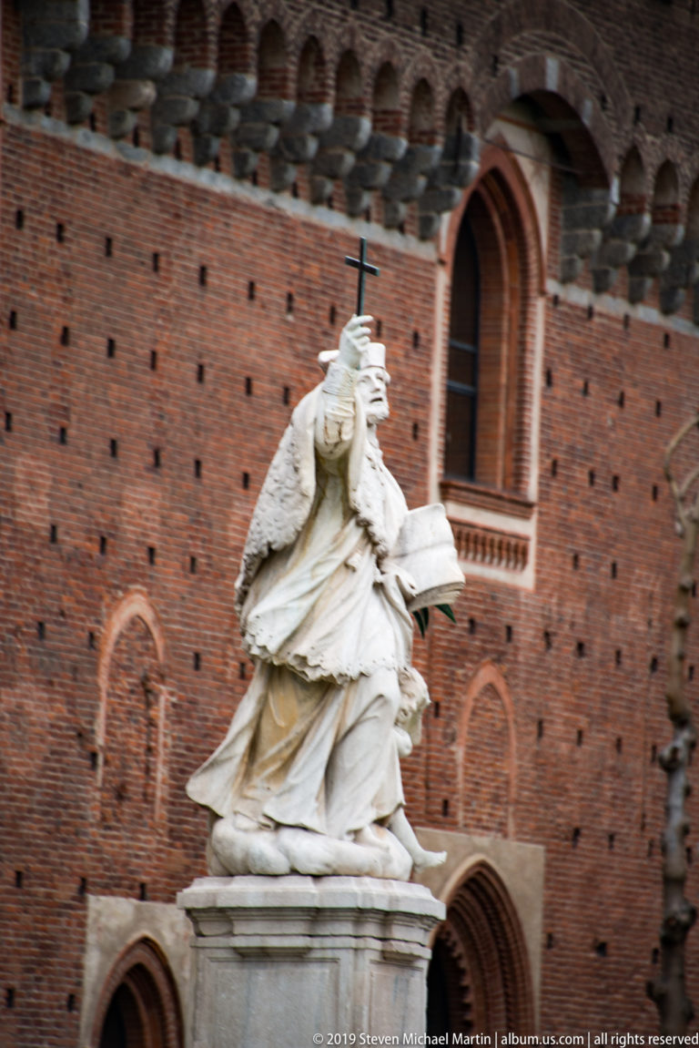 White marble seated female figure holding an open book against a red-brick wall with pointed-arch openings at Castello Sforzesco.