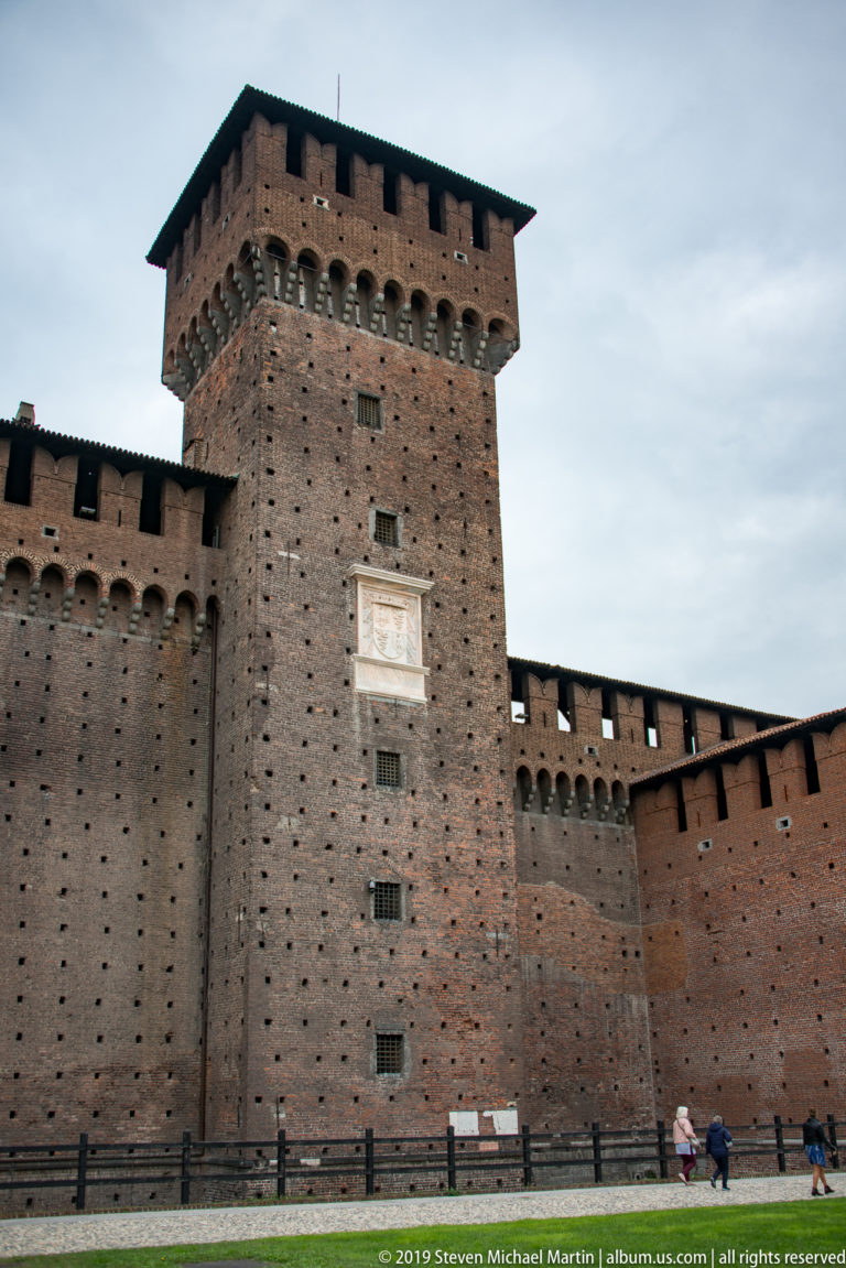 Massive square brick tower with projecting corbels, small openings, and a pale plaque along Castello Sforzesco’s perimeter wall.