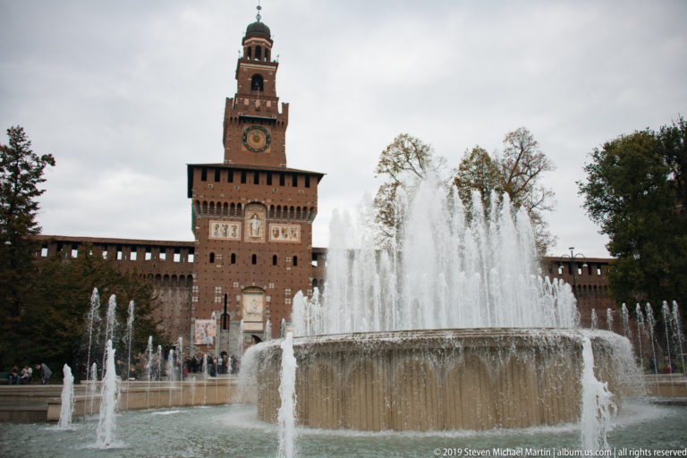 Large circular fountain centered before the Torre del Filarete, with castle walls extending behind under a cloudy sky.