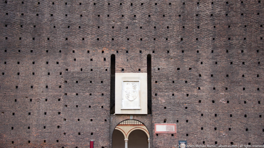 Pale heraldic plaque above a small arched doorway in a broad red-brick wall punctured by rows of square holes.