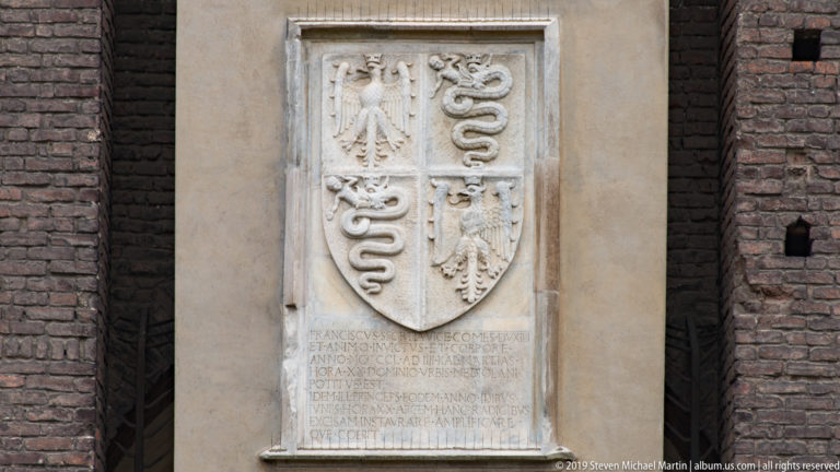 Pale carved stone plaque with quartered eagles and serpents above a Latin inscription, mounted on a dark brick wall.