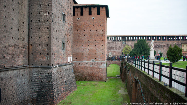 Square brick tower beside the grassy moat and an arched pedestrian bridge at Castello Sforzesco in Milan.