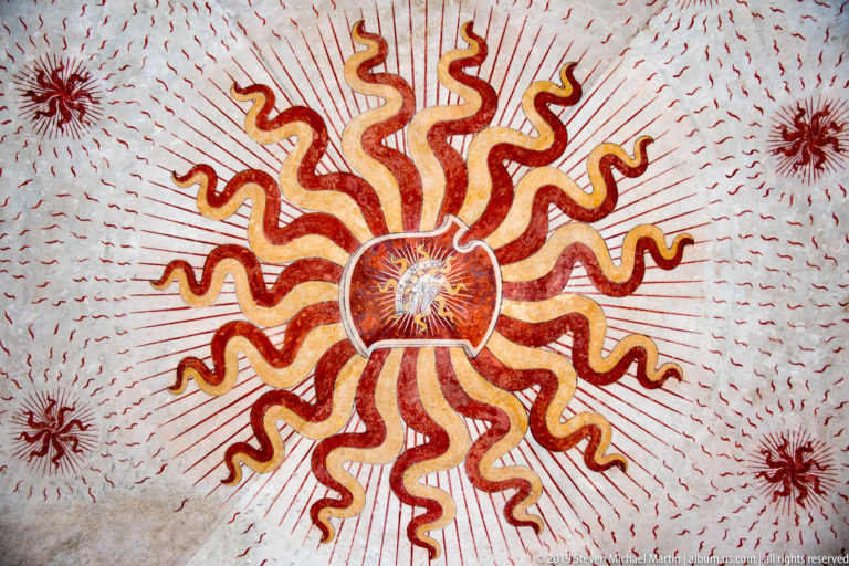 Close view of a circular red-and-ochre sunburst medallion with curling rays and fine radiating strokes on a pale vault.