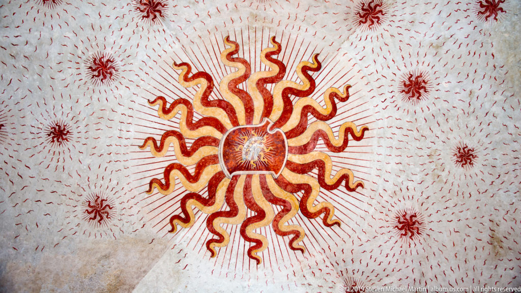 Large red-and-gold sunburst with flame-like rays and smaller radiant motifs painted across a pale vault at Castello Sforzesco.