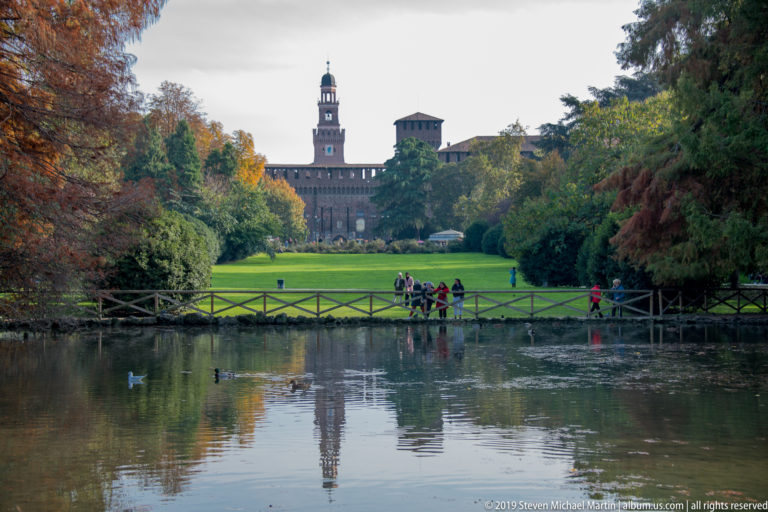 Castello Sforzesco and the Torre del Filarete beyond a reflective pond, green lawn, trees, and pedestrians in Parco Sempione.