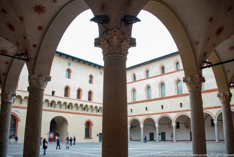 Large stone column and arches frame a pale inner courtyard with brick-trimmed windows at Castello Sforzesco.
