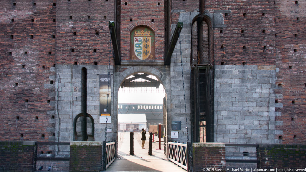 Pointed arched gateway through brick and stone, topped by a biscione shield and approached by a narrow railed bridge.