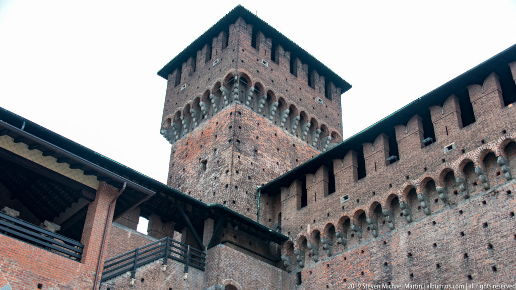 Tall square brick tower with projecting corbels and overhanging roof rising above covered galleries inside Castello Sforzesco.