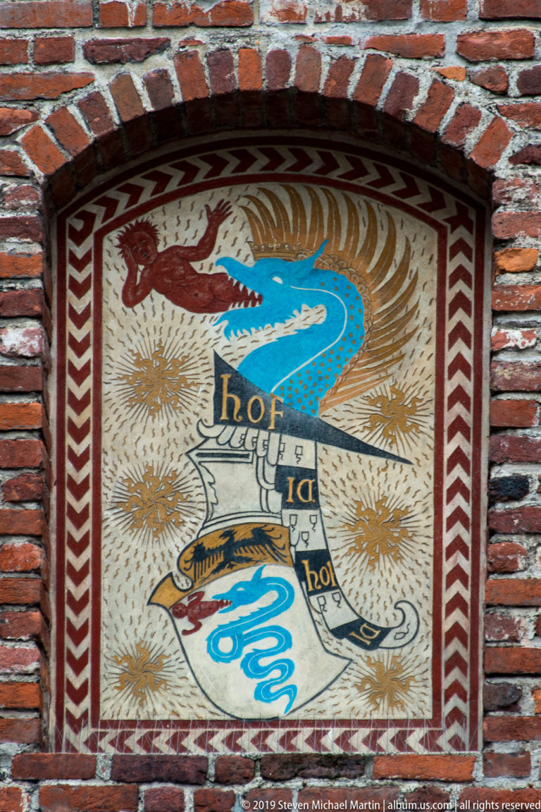 Painted heraldic panel in a brick recess showing a blue serpent, eagle, scroll, and red-and-white border at Castello Sforzesco.