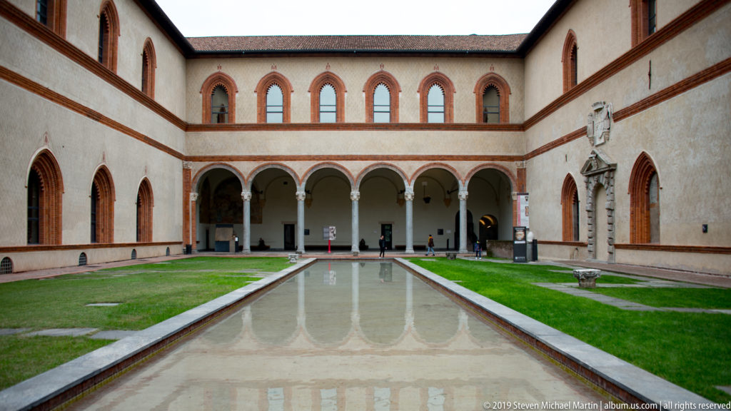 Long reflecting pool centered in a pale courtyard with brick-trimmed windows and a ground-floor arcade at Castello Sforzesco.