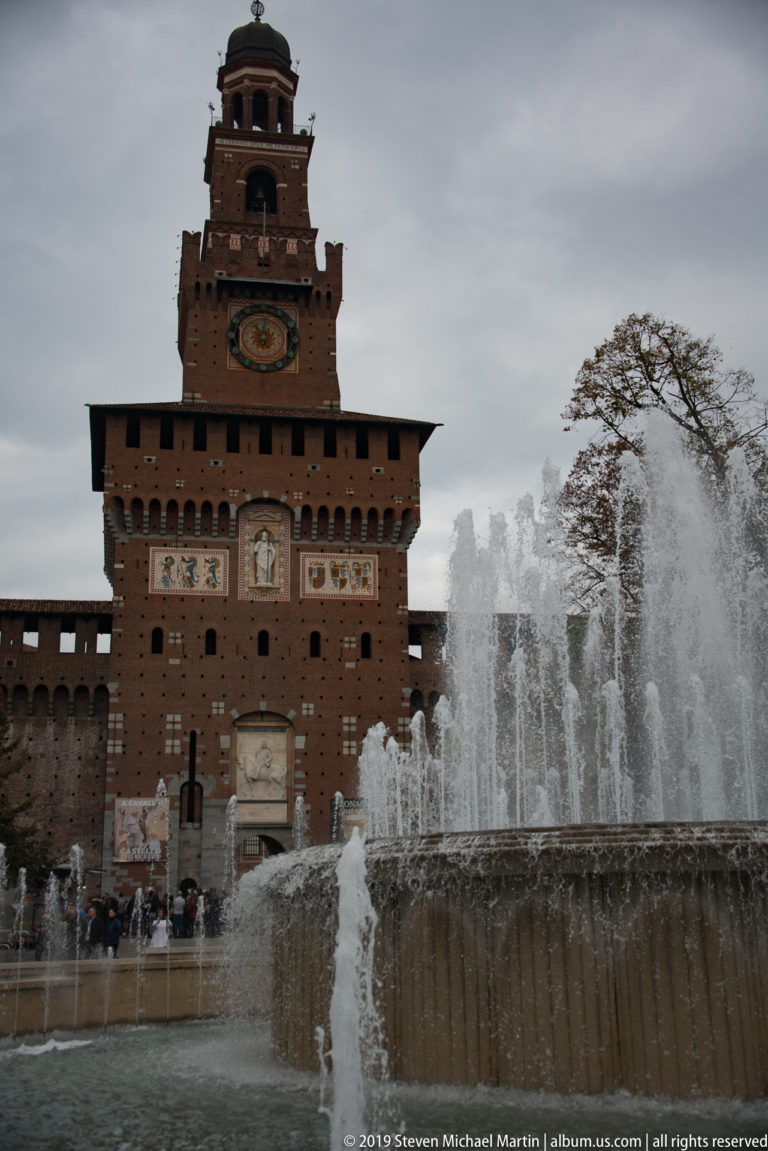 Fountain jets and basin in the foreground with the Torre del Filarete and Castello Sforzesco entrance behind.