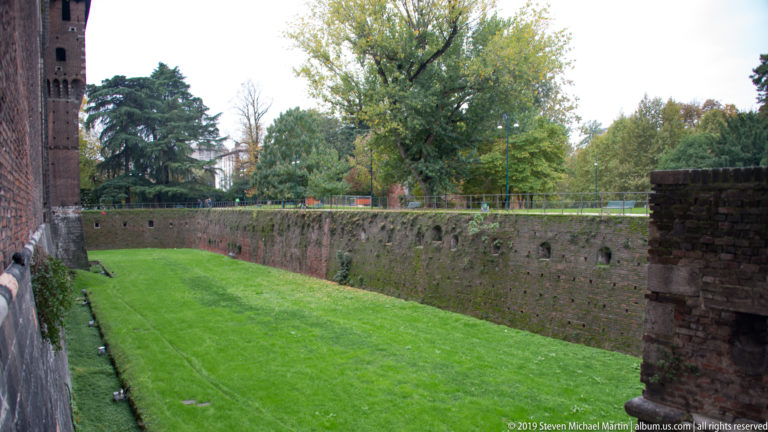 Long grassy dry moat enclosed by brick walls and trees along the exterior of Castello Sforzesco in Milan.