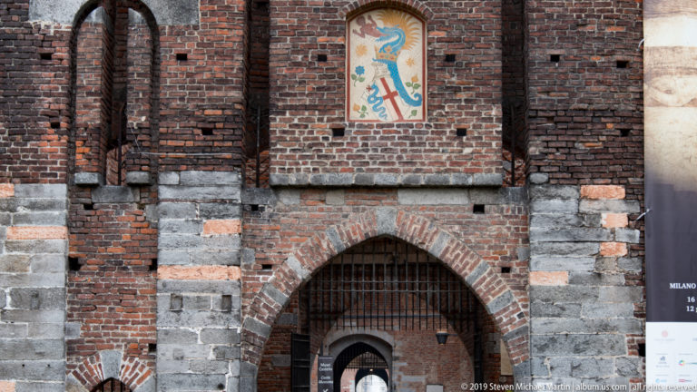 Painted heraldic panel with a blue serpent above an arched brick-and-stone gateway at Castello Sforzesco.