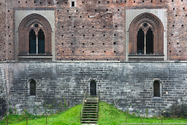 Two tall pointed-arch windows in a brick wall above a dark stone base, small lower openings, steps, and grassy moat.