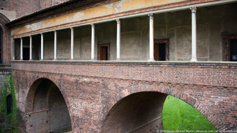 Two brick arches support a raised open loggia with slender columns above the grassy moat at Castello Sforzesco.