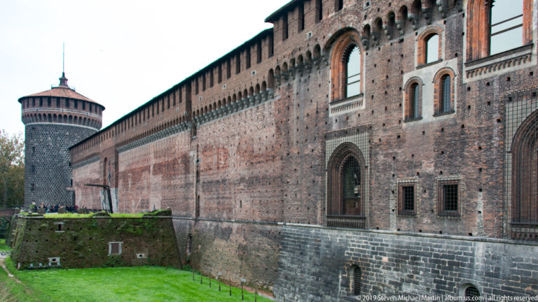 Long brick exterior wall of Castello Sforzesco receding toward a round corner tower beside the grassy moat.