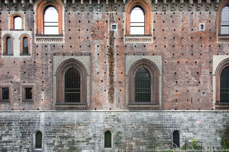 Pointed-arch grilled windows set in red brick above a dark stone base on an exterior wall of Castello Sforzesco.